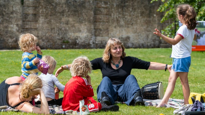 A woman is sitting on the ground with her arms stretched open wide. She has a group of children sitting and listening to her. It is a sunny day with blue sky.